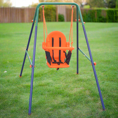 A swing set in the grass in a fenced yard for a toddler with an orange chair and safety harness. Steel frame with green detail along the top quarter. 